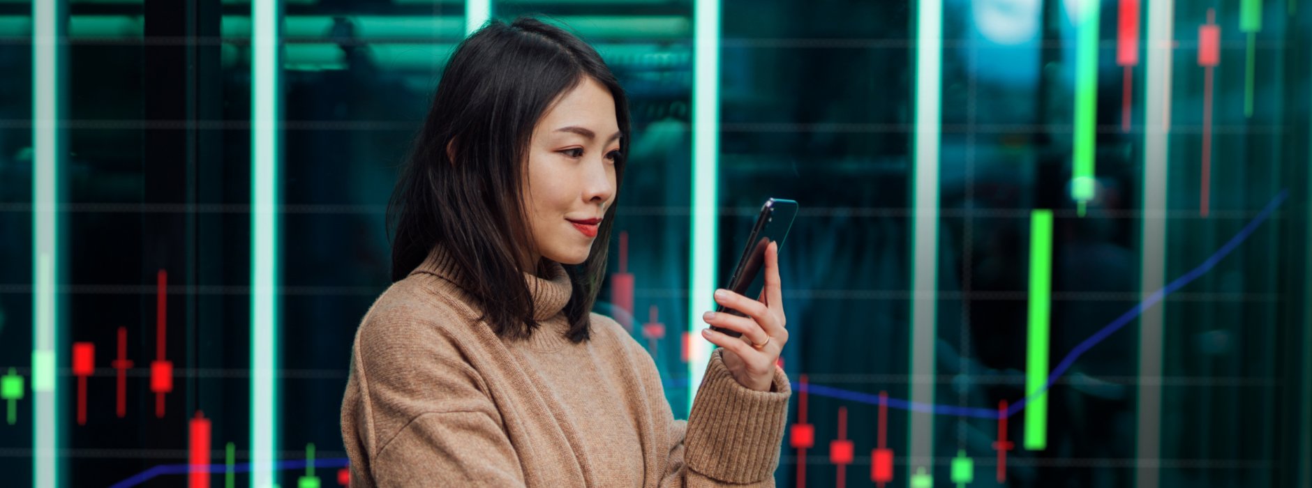 Asian woman looking at her phone standing in front of a background showing a trading chart