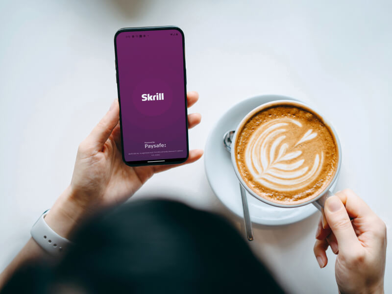 Person opening the Skrill mobile app while enjoying a coffee